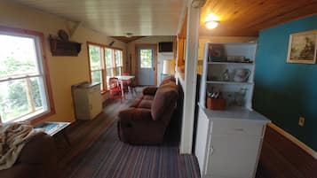 A cozy cabin interior with a sofa, a small table, a kitchenette, and a window overlooking greenery.