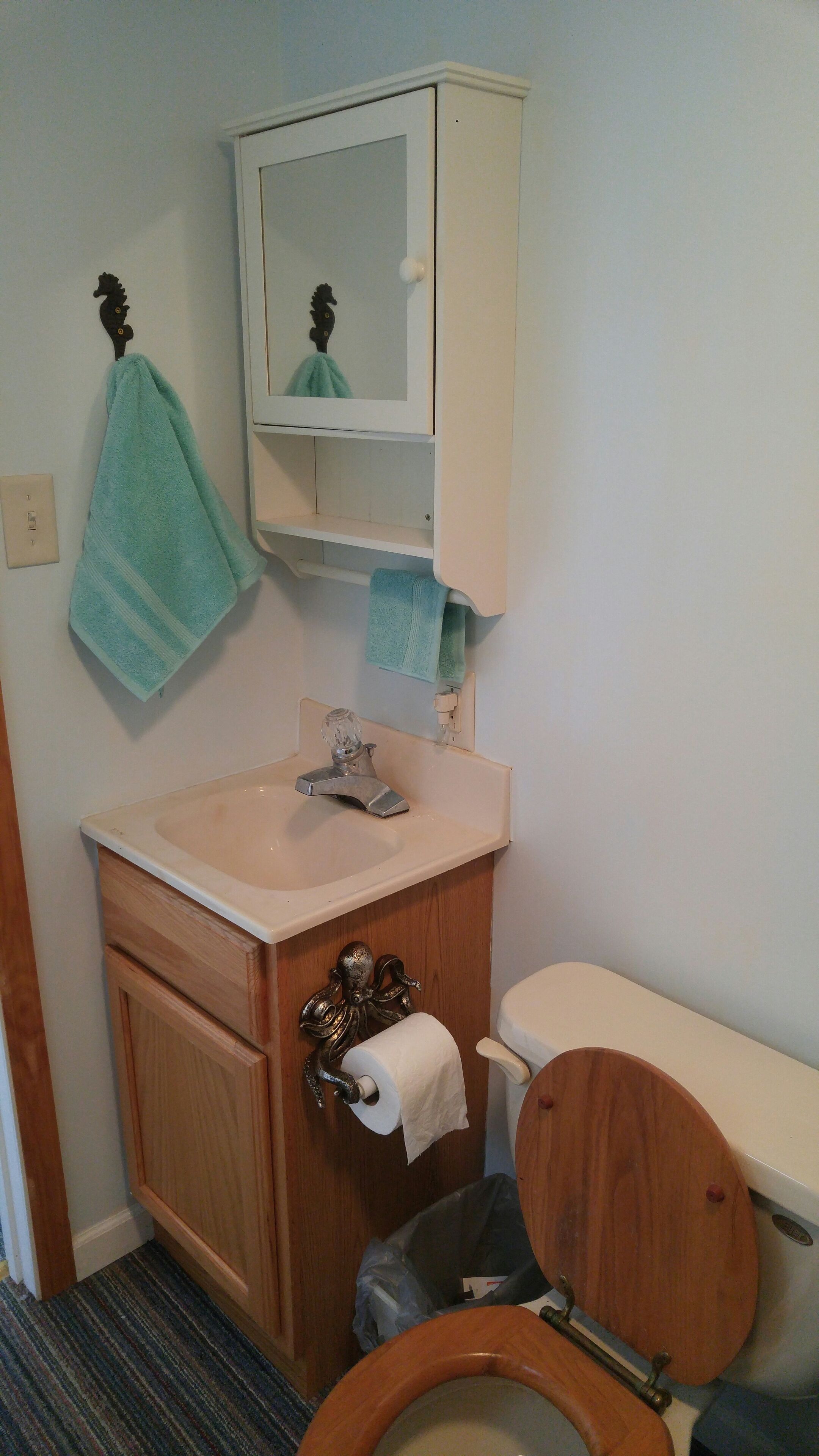 A bathroom with a wooden toilet, a white sink with a glass soap dispenser, a mirror above the sink, and a towel hanging on a hook.