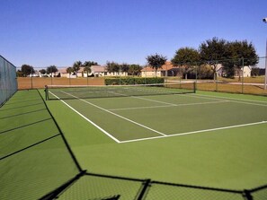 A tennis court with a green surface and white boundary lines, surrounded by a fence and trees.