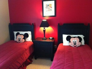 Une chambre avec deux lits, chacun agrémenté d’un coussin à l’effigie de Mickey Mouse, une table de chevet avec une lampe et un tableau encadré au mur.