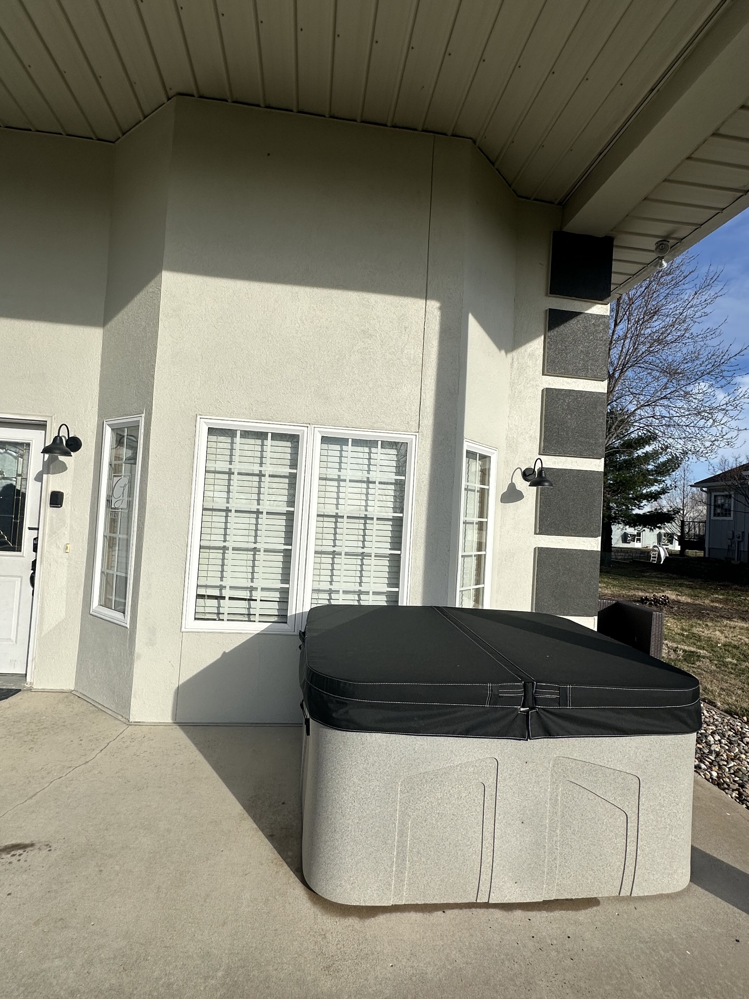A large, rectangular, beige-colored mattress placed outside a house with a sloped roof and a window with blinds.
