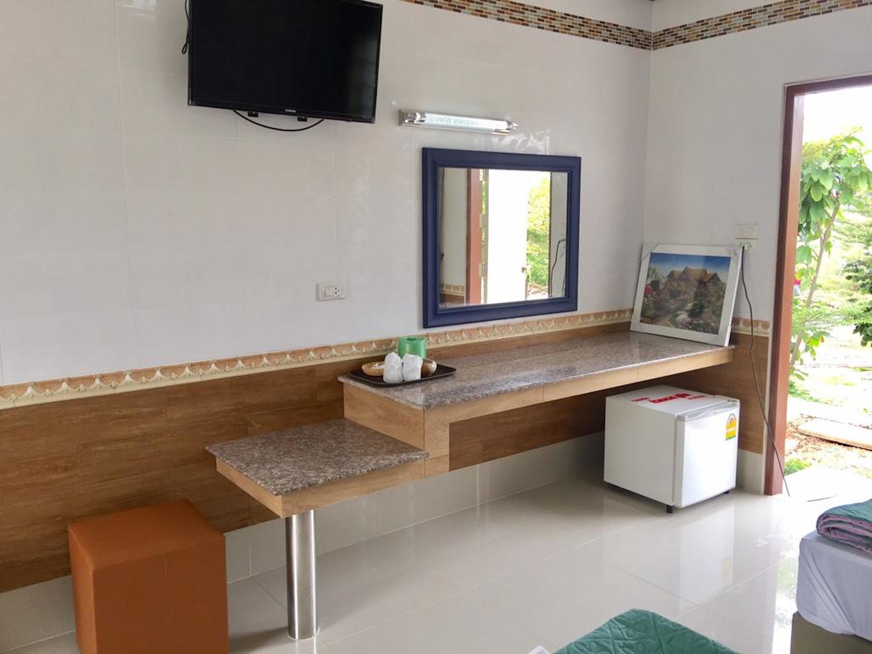 A hotel room with a TV mounted on the wall, a small refrigerator, a wooden bench, and a window with a view of greenery.