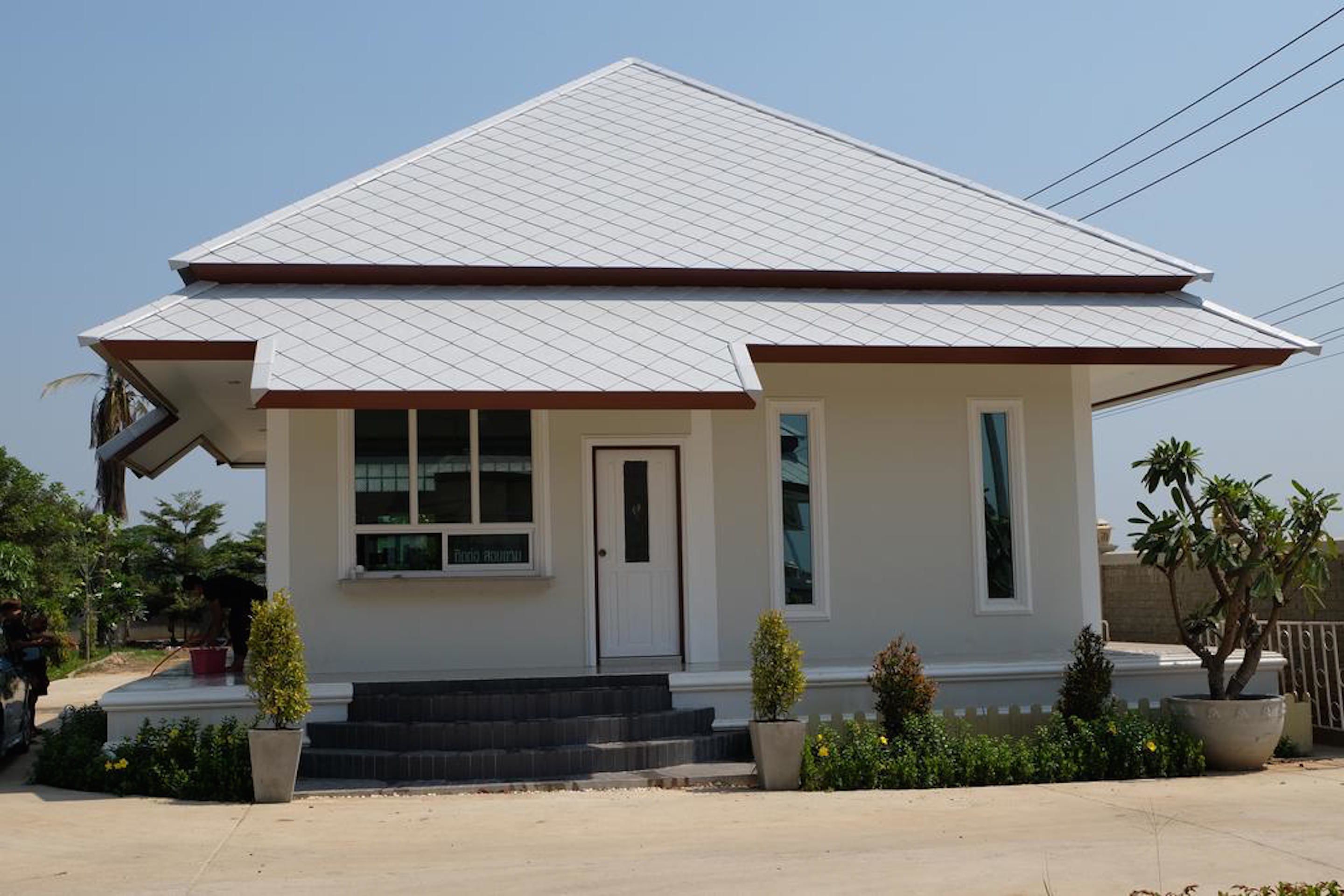 A small, single-story house with a white exterior and a brown roof. It features a front door, a small porch with steps, and a potted plant on the porch.