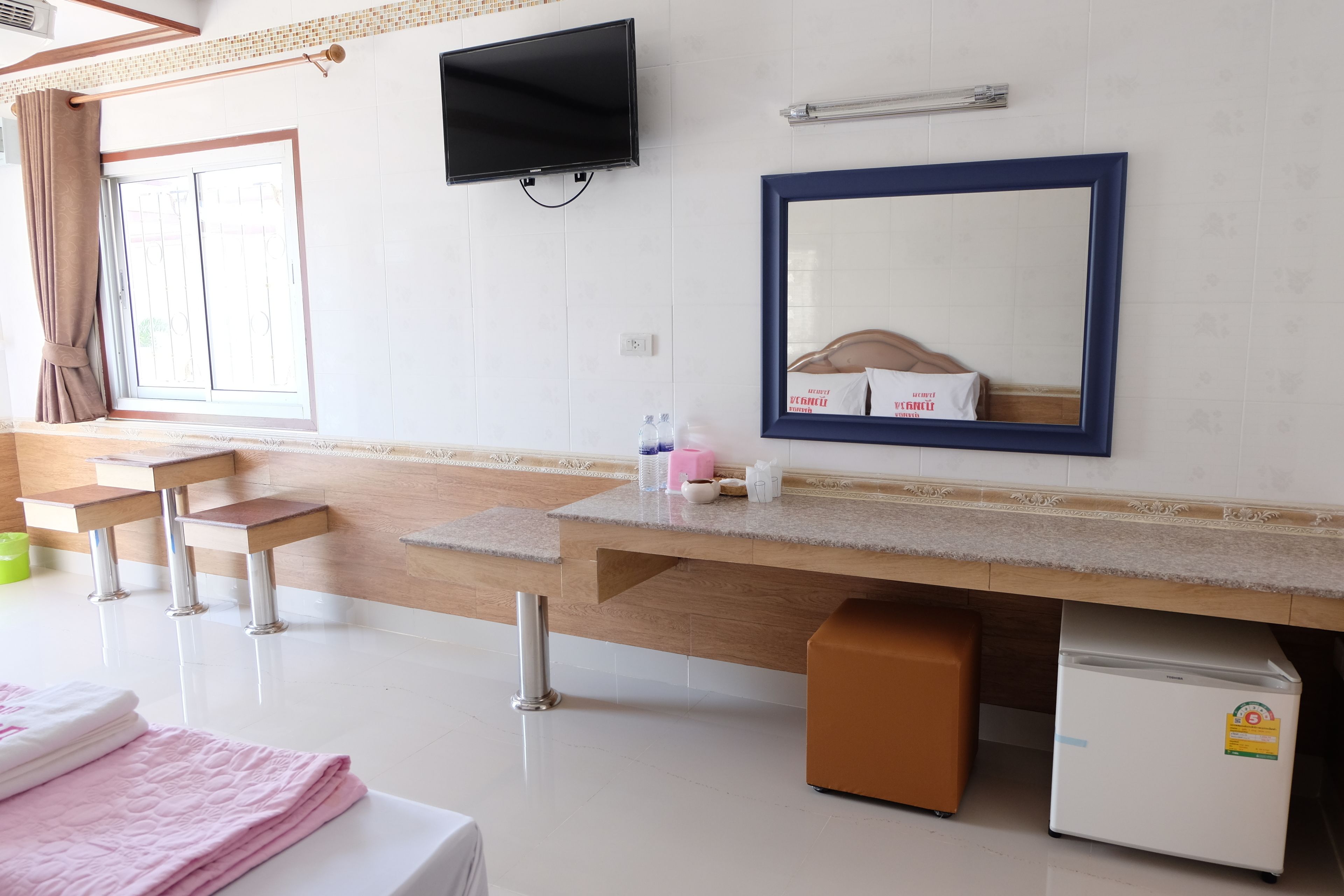 A hotel room with a wooden vanity, a flat-screen TV, a mirror, a small refrigerator, and a window with curtains.