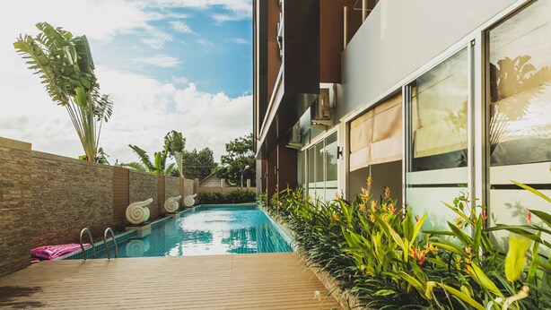 A swimming pool with a wooden deck, surrounded by plants and a stone wall. - Condo in Karon in Chic Condo - Unit A609 (Karon)
