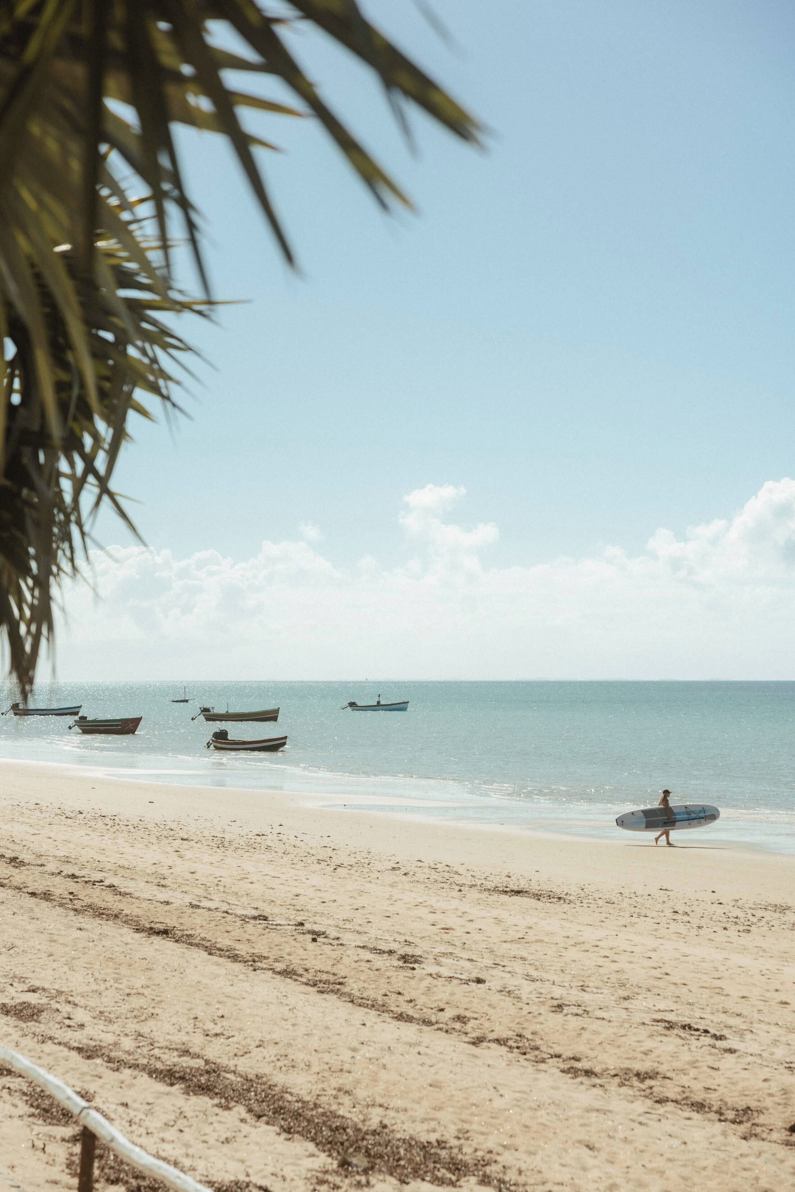 A beach with boats and a person carrying a surfboard.
