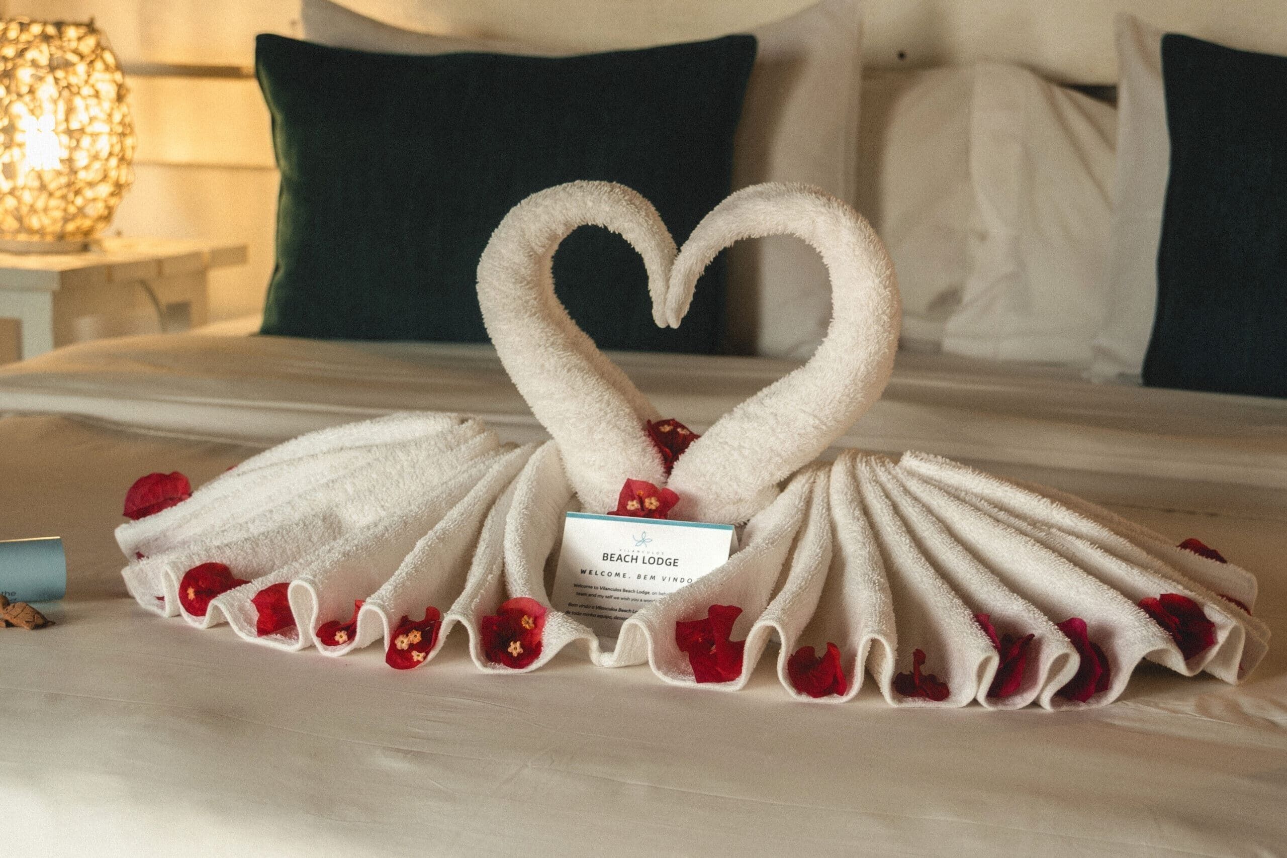 A neatly made bed with a towel arrangement in the shape of a heart, a welcome card, and scattered rose petals.