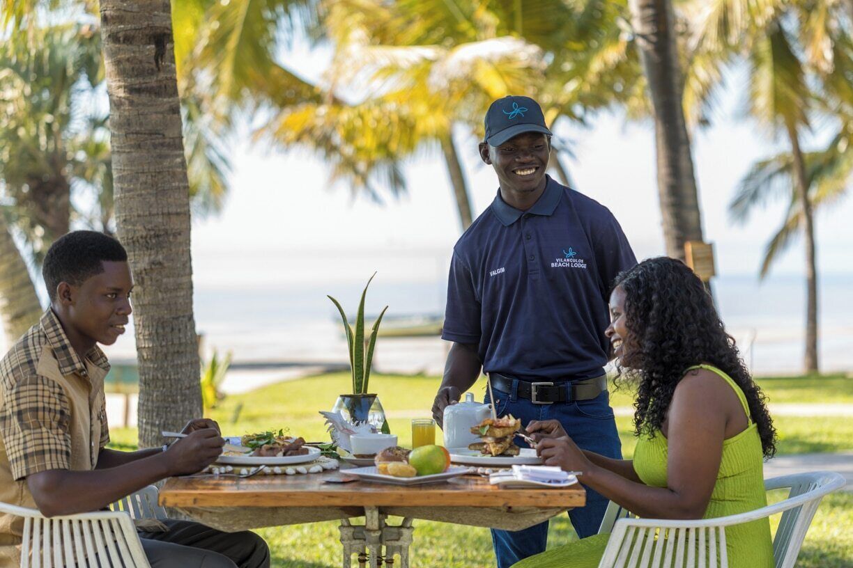 Un grupo de personas cenando al aire libre, con una mesa dispuesta con comida y bebidas, palmeras y vista al océano de fondo.