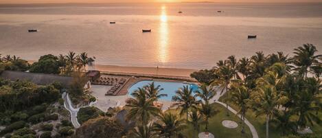 A resort with a swimming pool, palm trees, and a view of the ocean at sunset.
