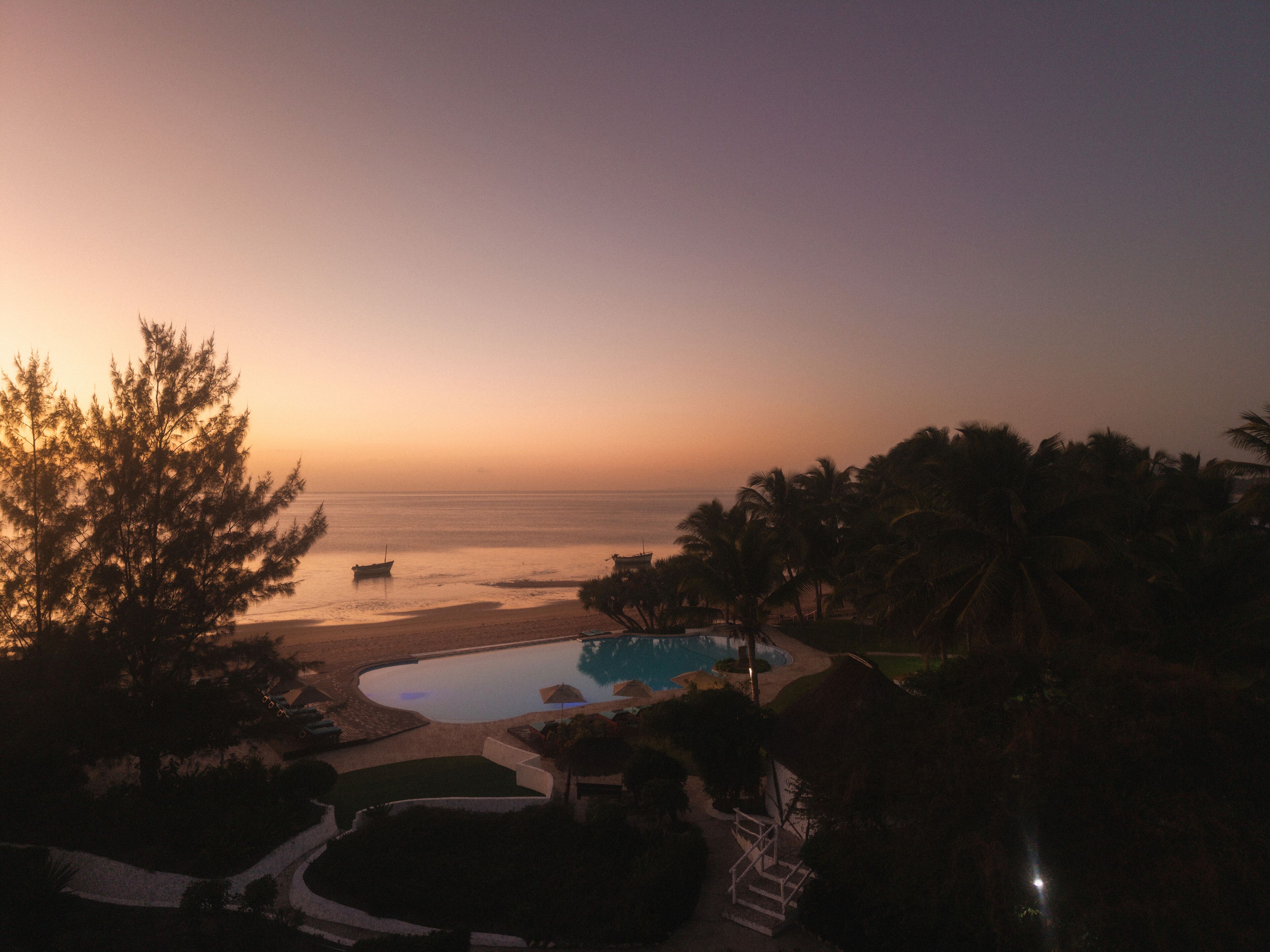 A resort with a pool, palm trees, and a view of the ocean at sunset.