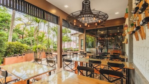 An indoor dining area with glass tables and chairs, a hanging light fixture, and a view of greenery outside.