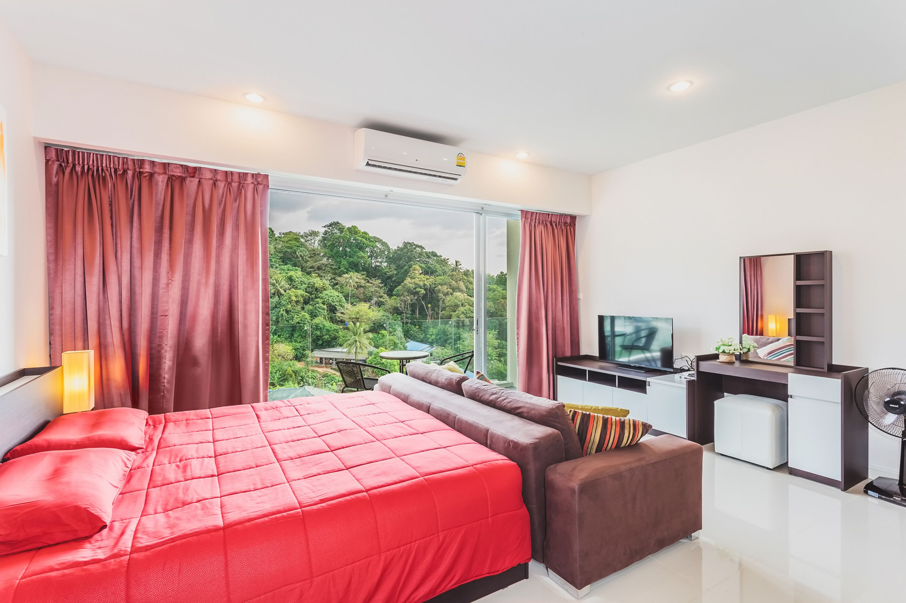 A hotel room with a large bed, a sofa, a desk with a TV, and a view of lush greenery.