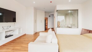 A modern hotel room with a large bed, a white sofa, a flat-screen TV, and a large window.