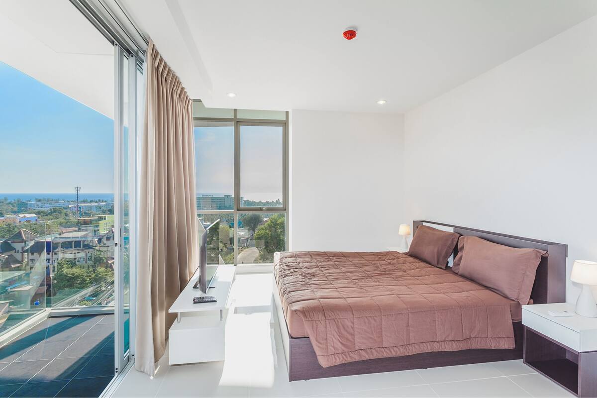 Un dormitorio moderno con una cama grande, una mesita de noche y vistas a la ciudad a través de ventanales de gran altura.