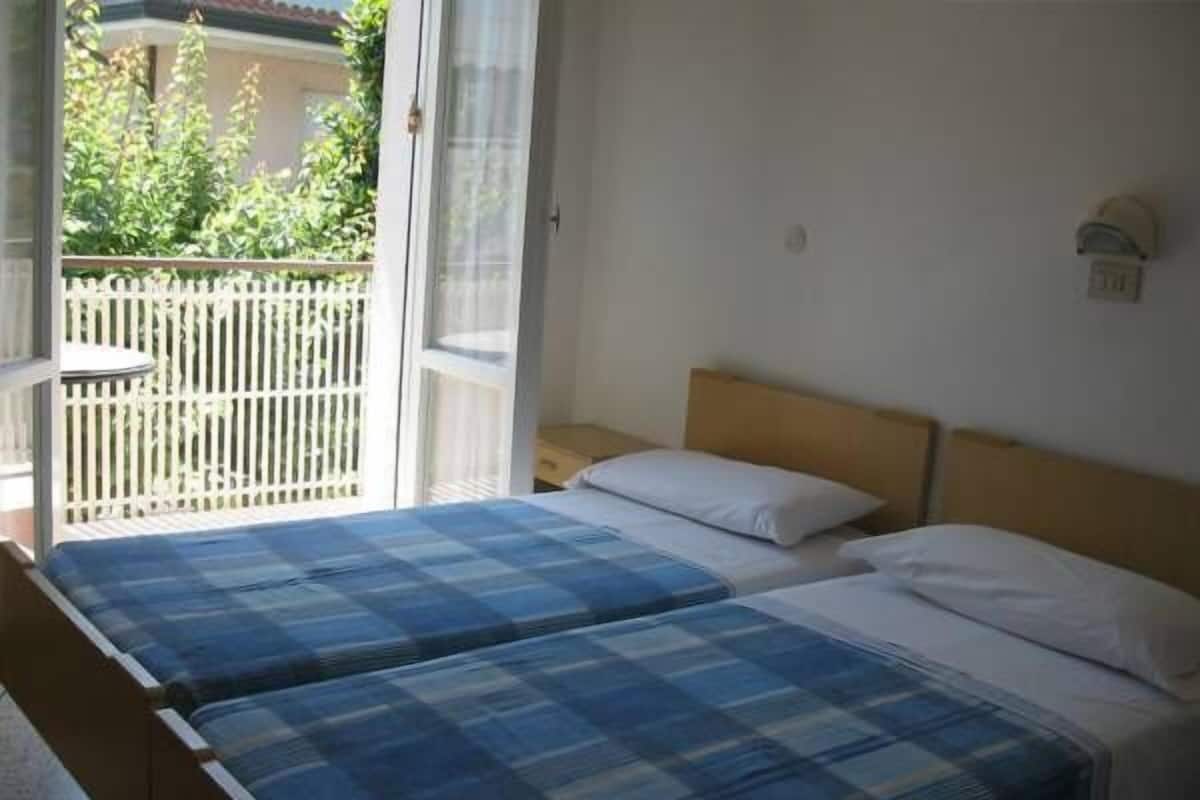 A room with two beds, a wooden headboard, a white wall, and a view of a balcony with a white fence and greenery.