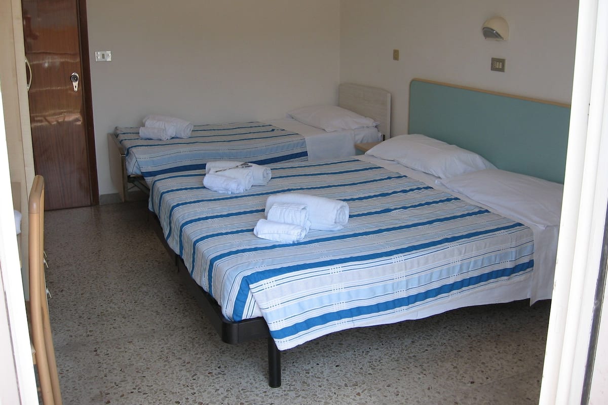 A hotel room with three beds, each with a striped bedspread and white pillows. There is a wooden door and a light switch on the wall.