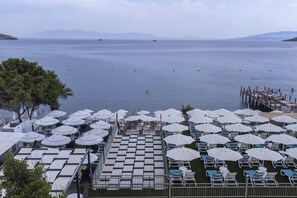 Terraza frente al mar con filas de tumbonas y sombrillas, con vista a un mar tranquilo y barcos a la distancia.
