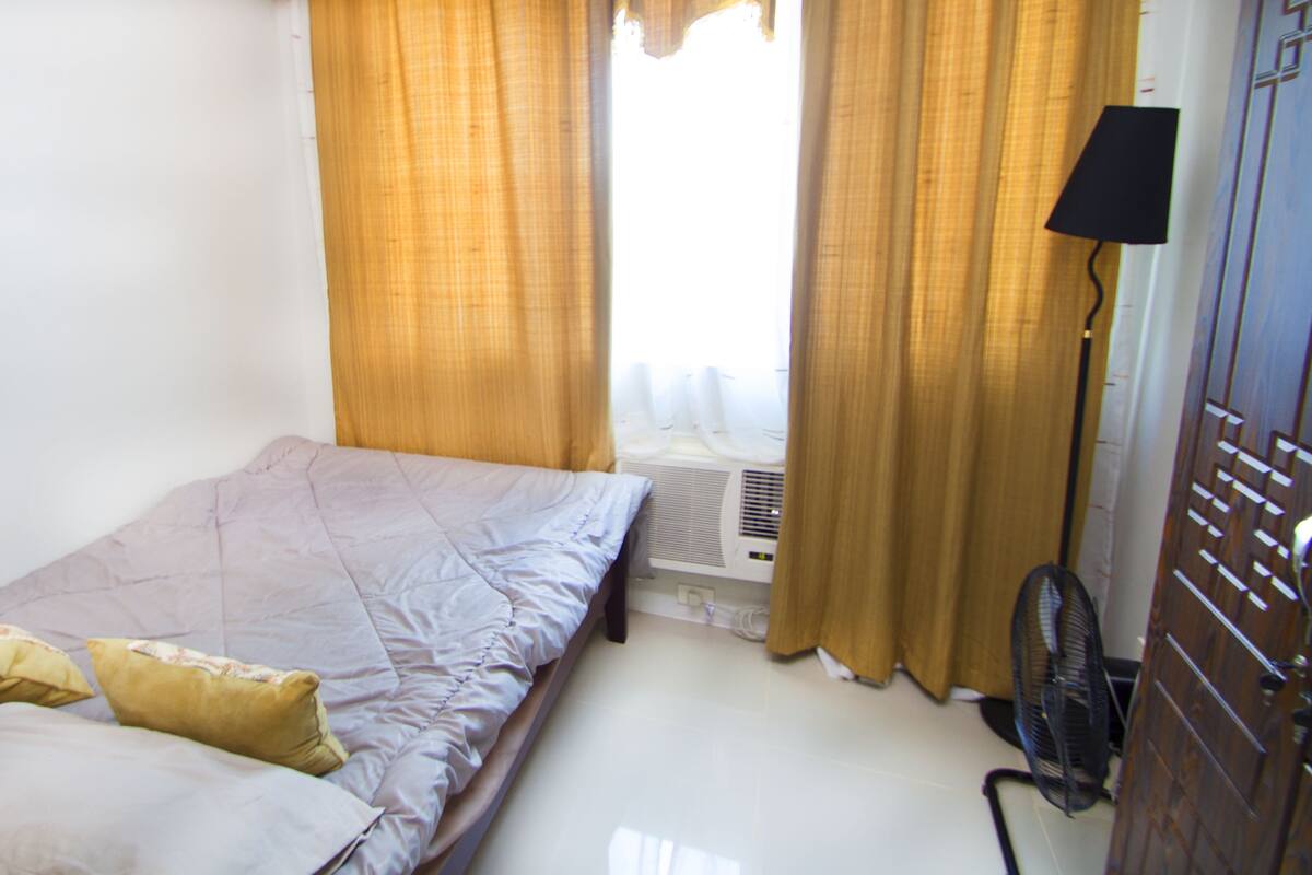 Un lit simple avec une couette de couleur claire et deux oreillers, une table de chevet avec une lampe et une fenêtre avec des rideaux.