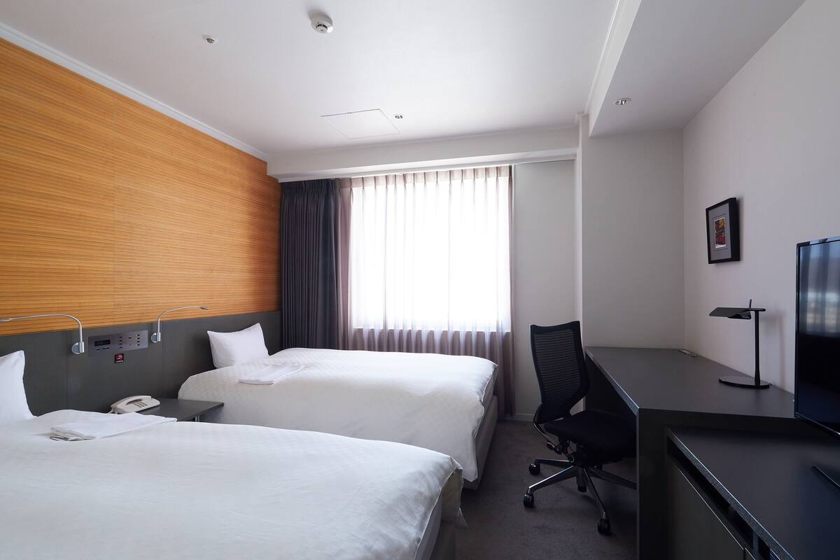 Hotelkamer met twee bedden, een bureau, een stoel, een televisie en een raam met gordijnen.