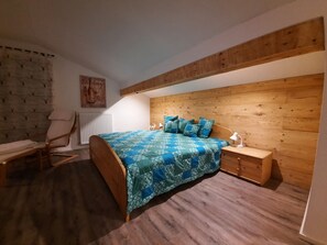 Une chambre avec une tête de lit en bois, un lit recouvert d’une couvre-lit à motifs bleus, une table de chevet en bois avec une lampe, et une chaise avec une serviette.