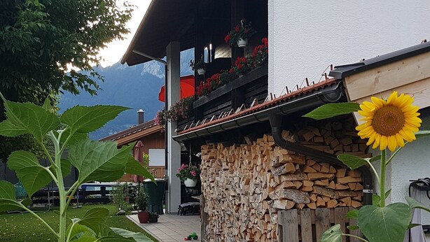 Una construcción rústica con fachada de madera, un balcón con flores rojas y un girasol grande en primer plano.