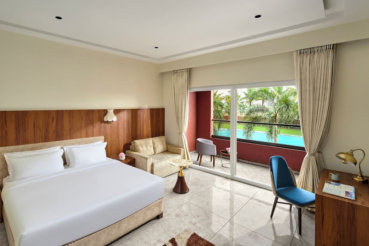 A hotel room with a large bed, a sofa, a desk with a chair, a small table, and a view of a swimming pool.