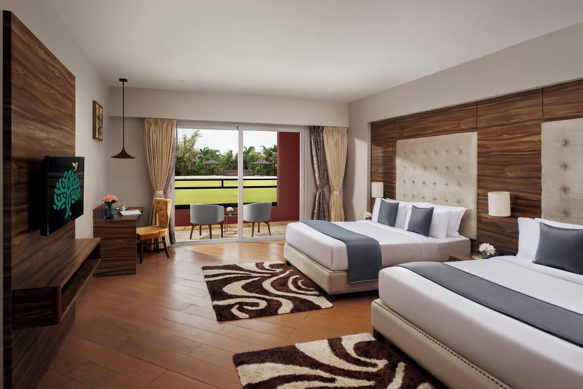 A modern hotel room with a large bed, a desk with a chair, a television, and a view of a green outdoor area.