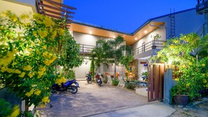 A courtyard with a motorcycle parked in the center, surrounded by greenery and a building with a wooden ceiling and a balcony. - Samsha Guesthouse (Hua Hin)