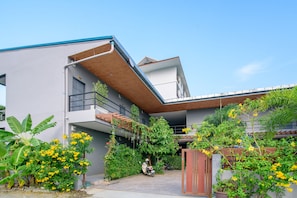 A modern two-story building with a balcony, surrounded by lush greenery and yellow flowers. - Samsha Guesthouse (Hua Hin)