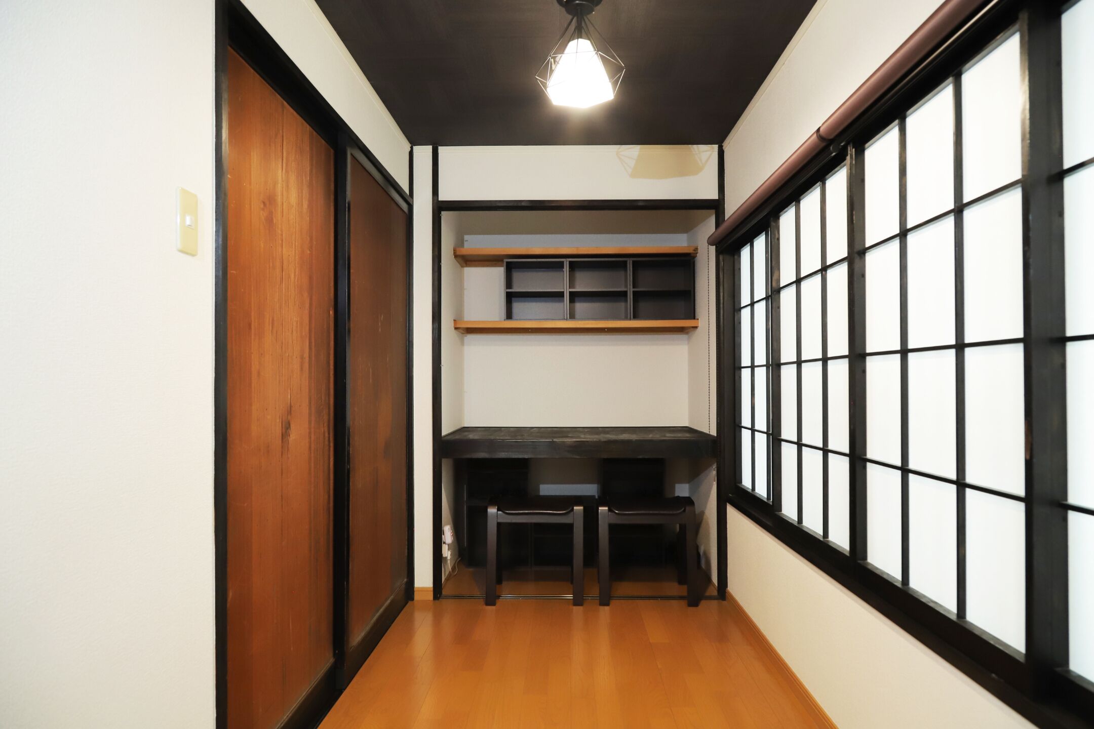 Un pasillo estrecho con suelo de madera, un escritorio con dos sillas y una ventana con diseño de cuadrícula.