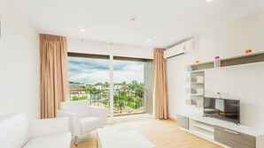 A modern living room with a white sofa, a white coffee table, and a TV on a white entertainment unit. Large windows offer a view of a balcony and greenery. - Condo in Kata in Ozonec- Unit D618 (Karon)