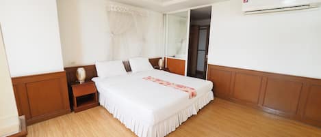 A hotel room with a large bed, wooden flooring, a nightstand, and an air conditioning unit.