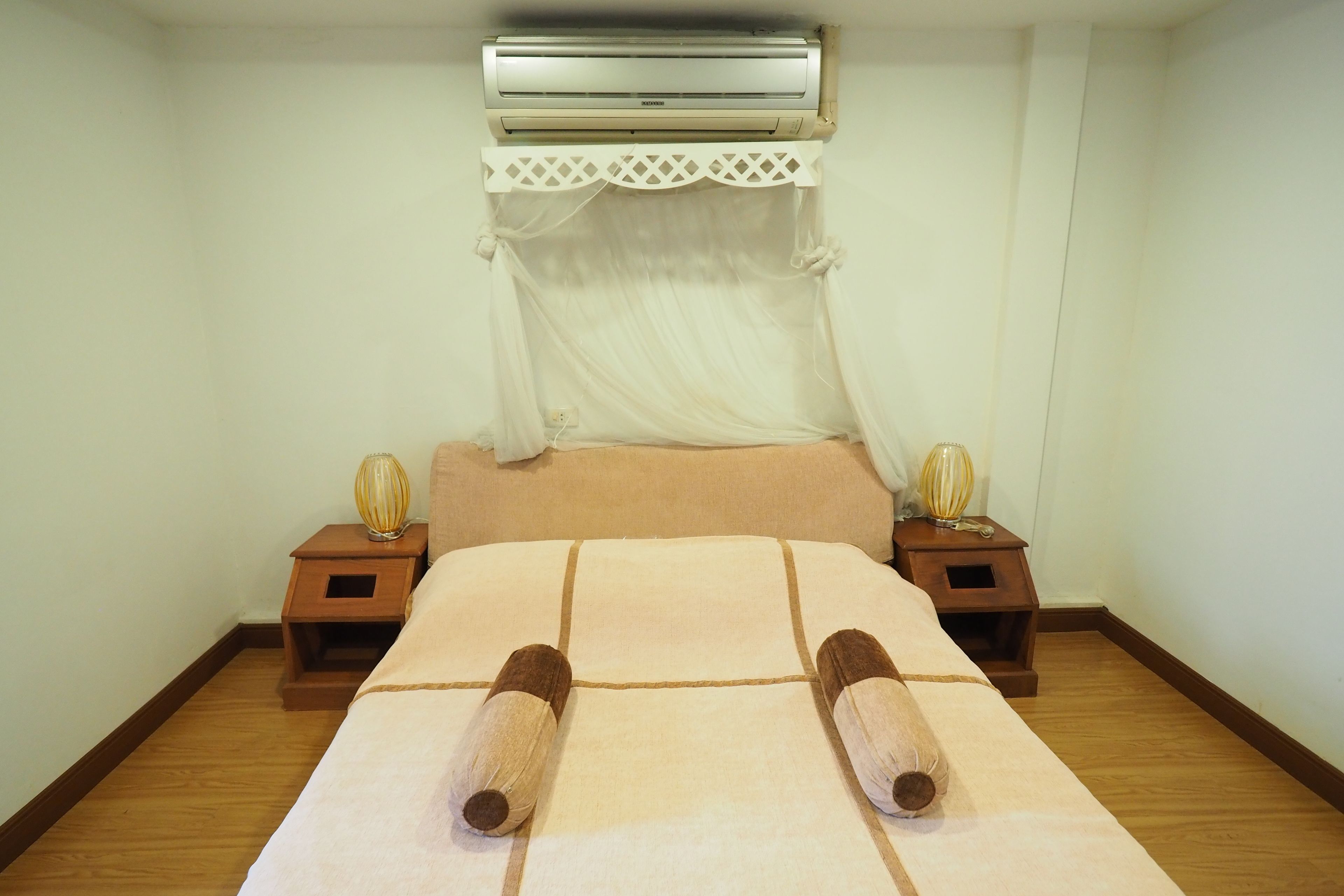 Een net opgemaakt bed met een witte dekbedovertrek en twee opgerolde handdoeken. Daarboven, boven het bed, hangt een witte airconditioningunit, en aan beide zijden staan nachtkastjes met lampen.