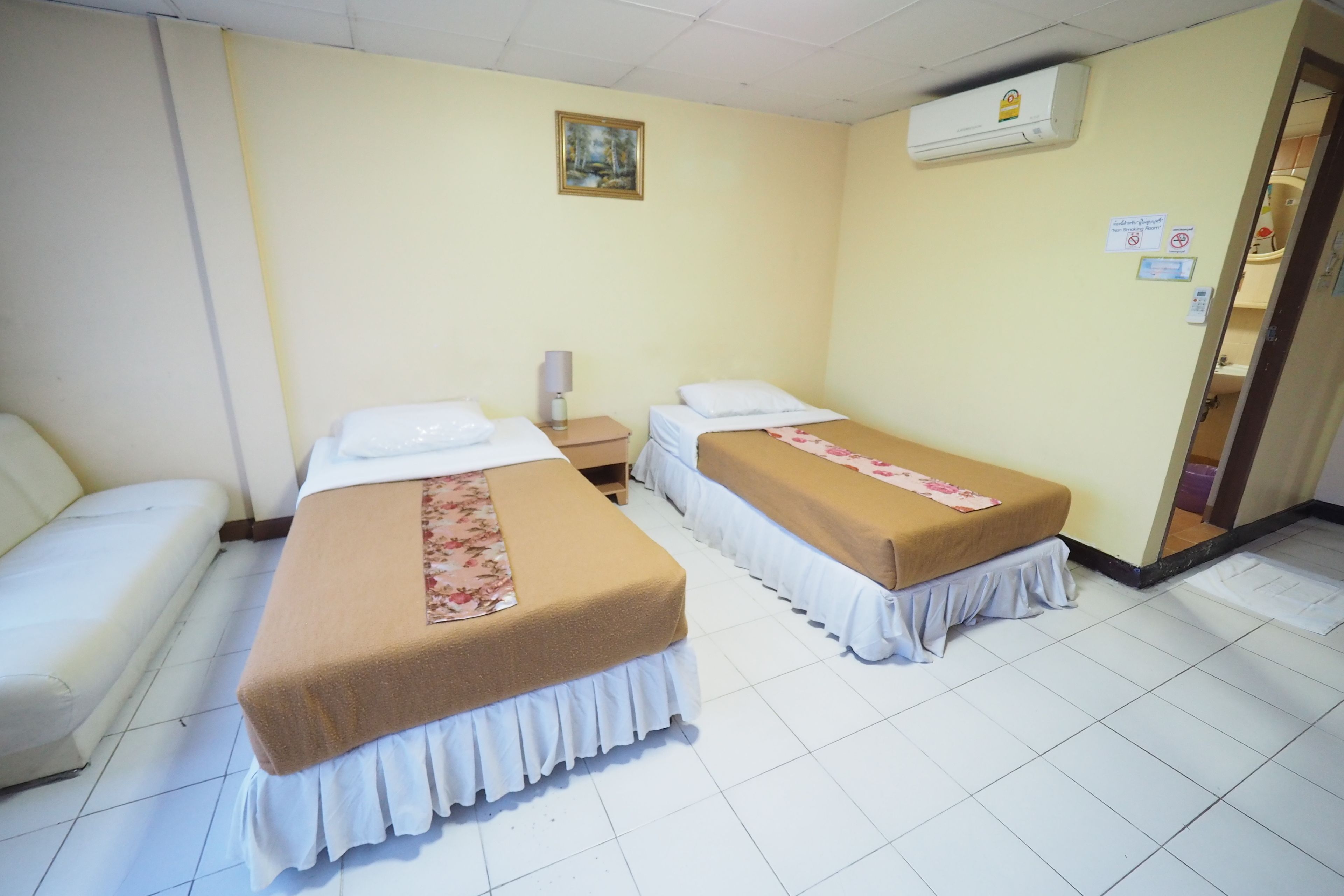 A hotel room with two beds, a sofa, a bedside table with a lamp, and an air conditioning unit.