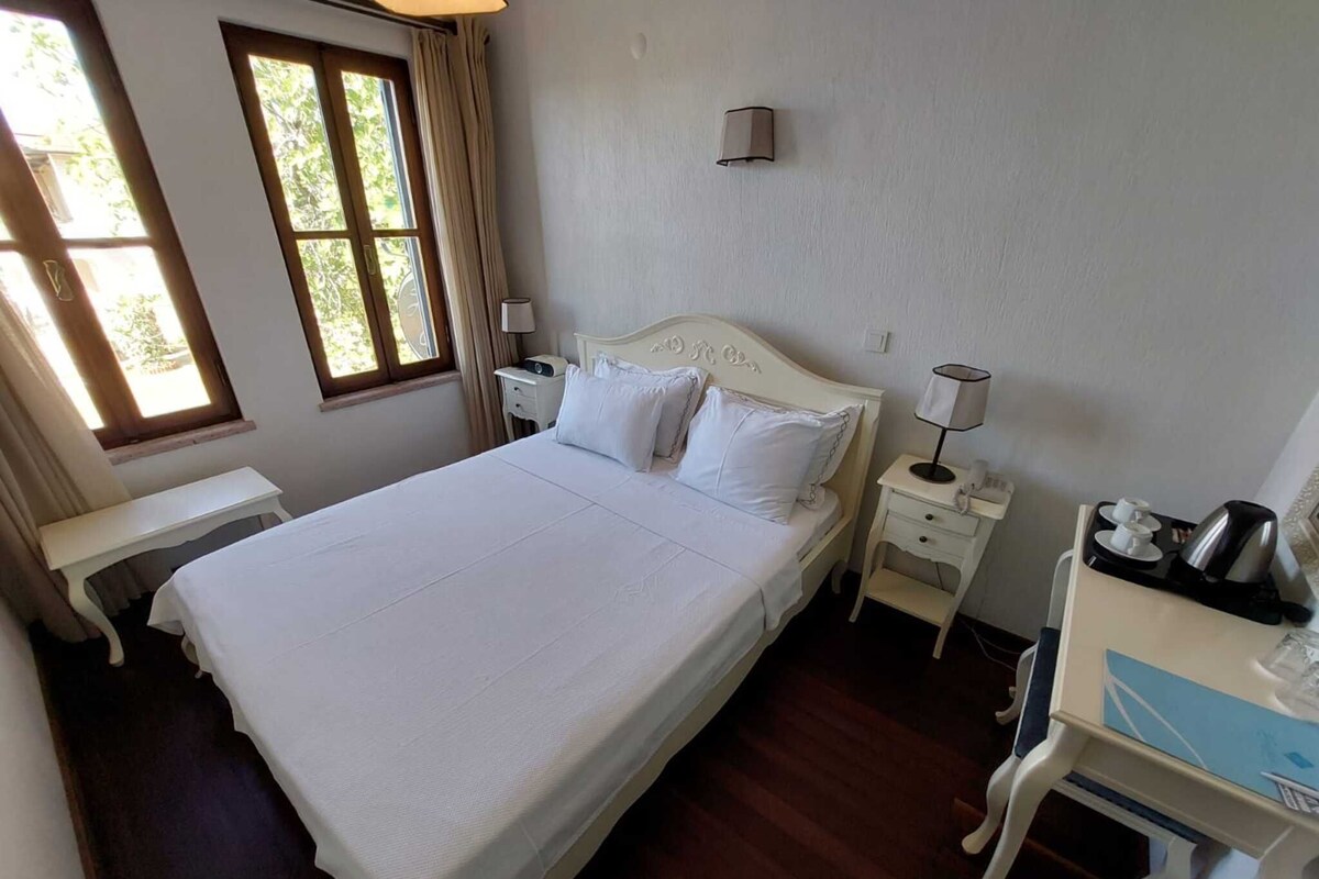 A neatly made bed in a room with a window, a bedside table with a lamp, and a desk with a coffee maker.