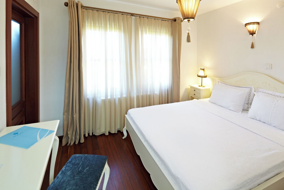 A hotel room with a large bed, a bedside table with a lamp, a desk with a chair, and a window with curtains.
