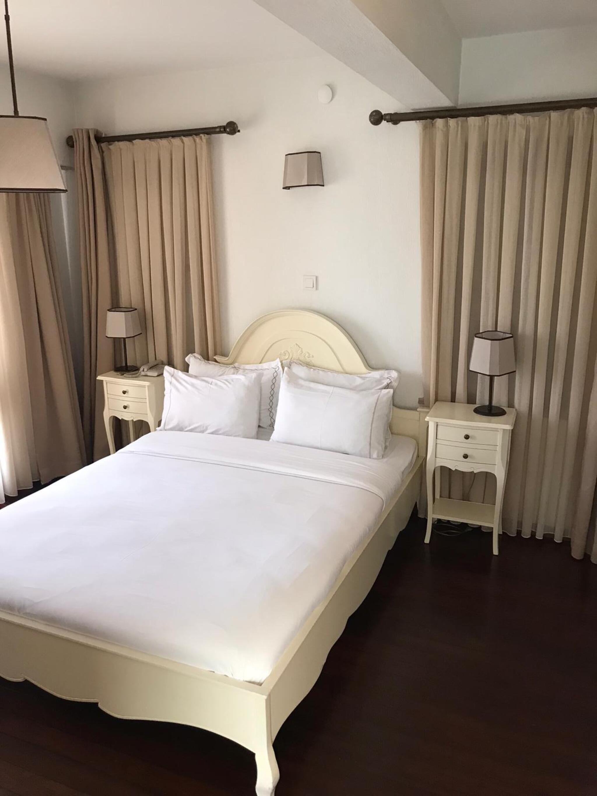 A neatly made bed with white linens, two bedside tables with lamps, and beige curtains.