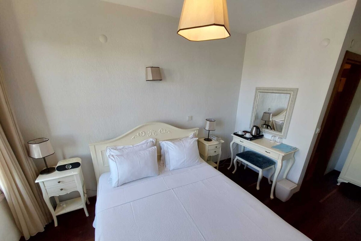 Habitación de hotel con cama, mesitas de noche, escritorio, silla, lámpara, espejo y ventana.