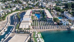 An aerial view of a coastal resort with a marina, a swimming pool, and a beach area with umbrellas.