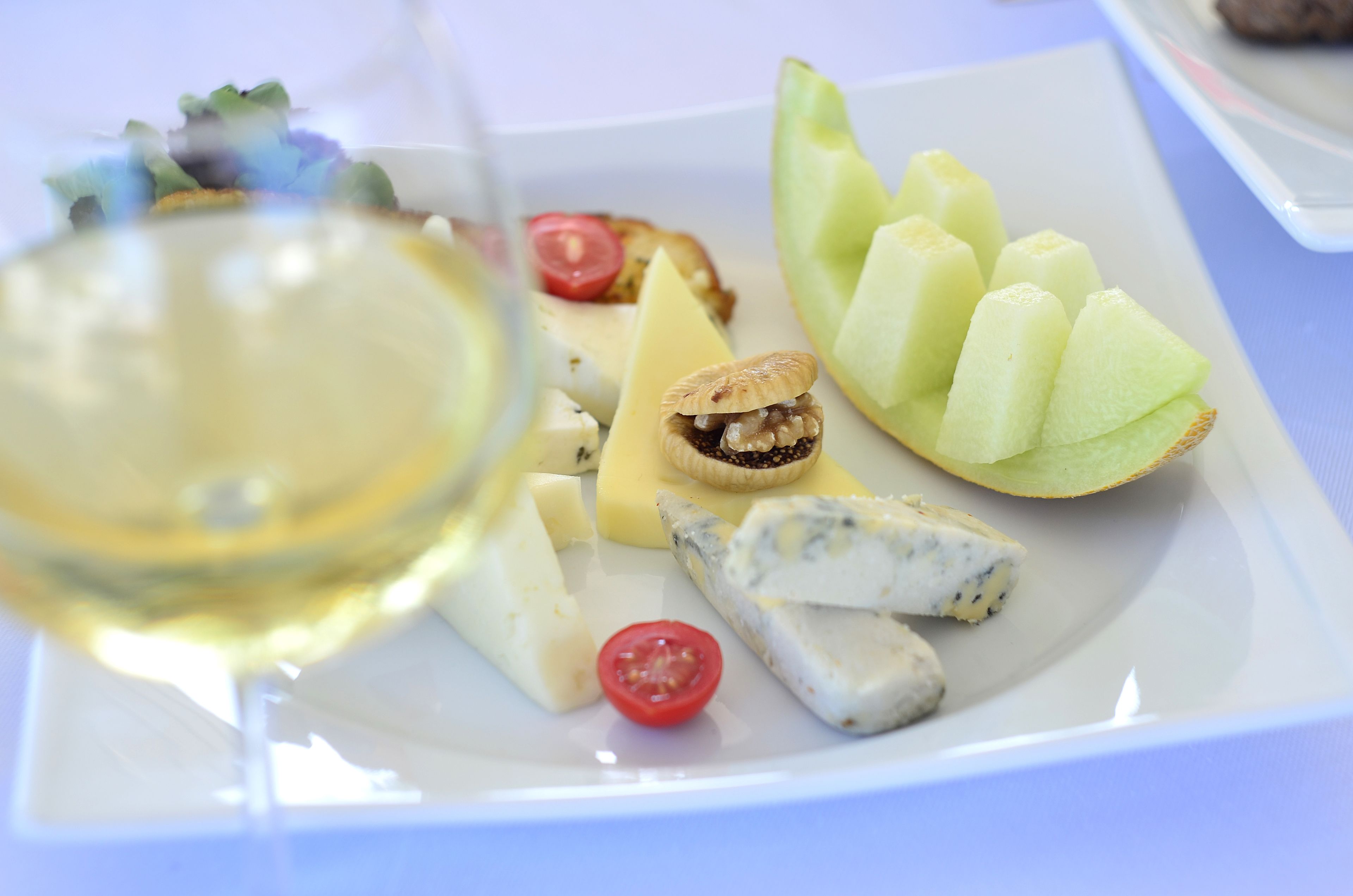 Une assiette de fromages et de fruits variés, comprenant une tranche de melon, une tomate cerise et un morceau de fromage bleu, accompagnée d’un verre de vin blanc.