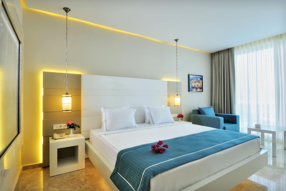 A modern hotel room with a large bed, bedside lamps, a small table, and a view of the city through the window.