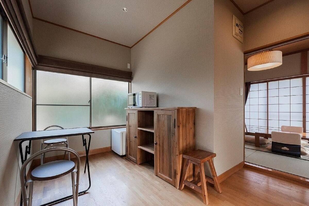 Ein kleines Zimmer mit einem Holzschrank, einem kleinen Hocker, einem Tisch mit zwei Stühlen und einem Fenster mit Blick.