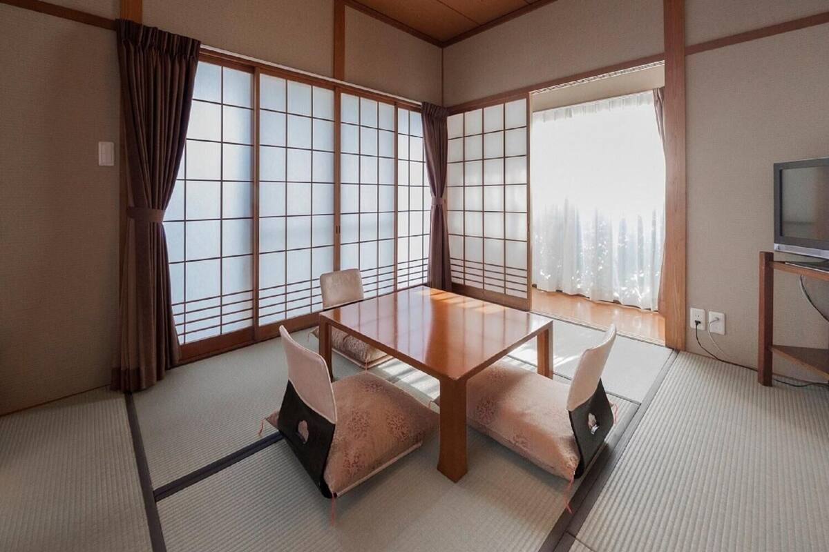 Ein traditioneller japanischer Raum mit Tatami-Boden, einem niedrigen Tisch, Kissen, verschiebbaren Shoji-Türen und einem Fernseher auf einem Holzständer.