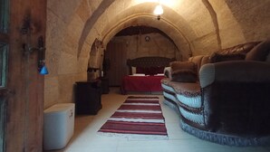 A cozy room with a wooden door, a red bedspread, a patterned rug, and a leather sofa. - Sandik Cave Hotel (Ürgüp)