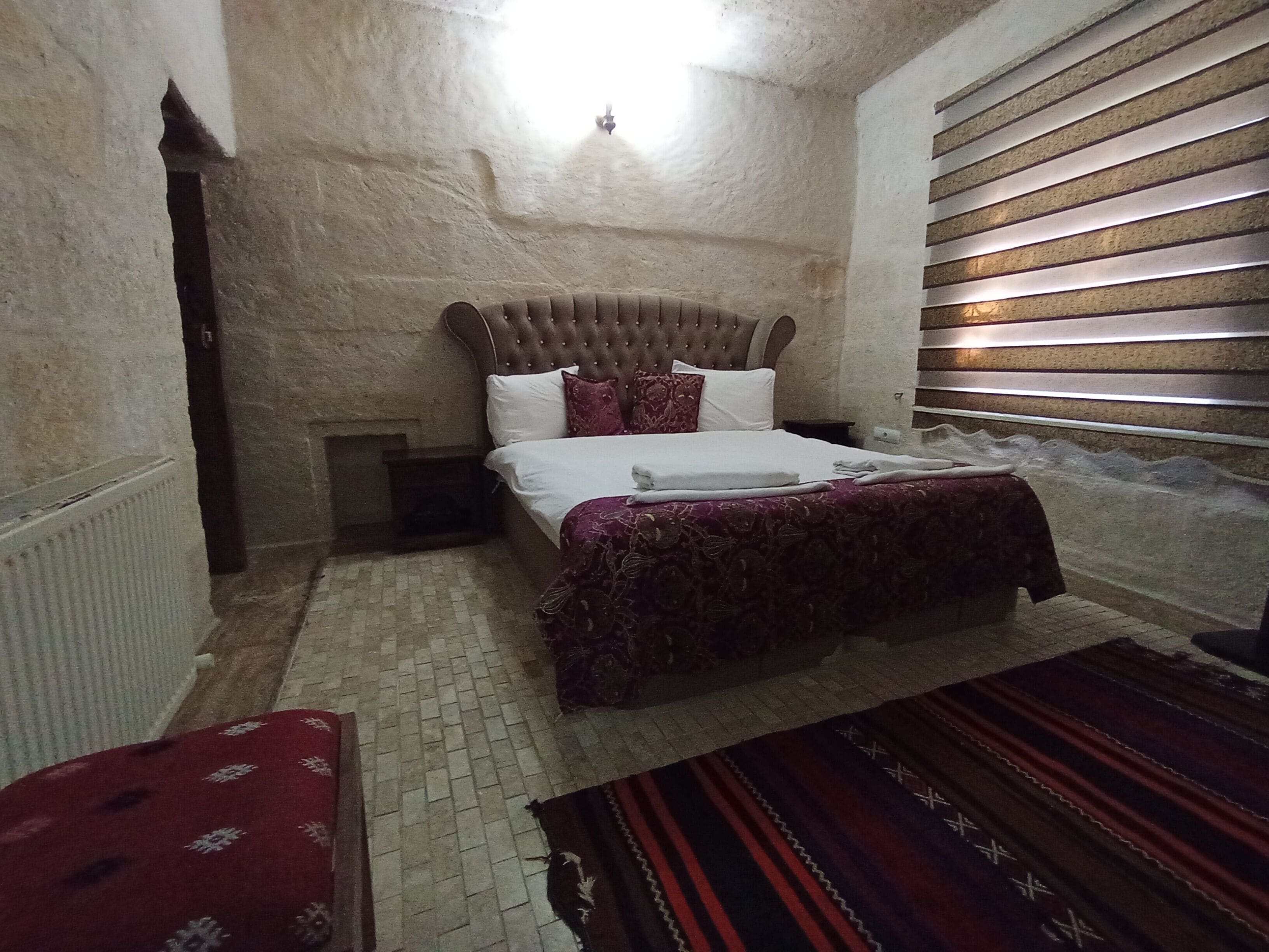 Un dormitorio con una cama grande, cabecera, mesita de noche, un banco rojo y una alfombra estampada.