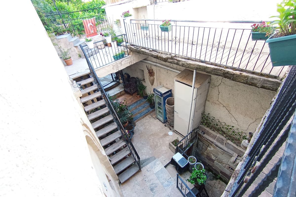 A courtyard with a wooden staircase leading to a balcony, surrounded by potted plants and a water dispenser.