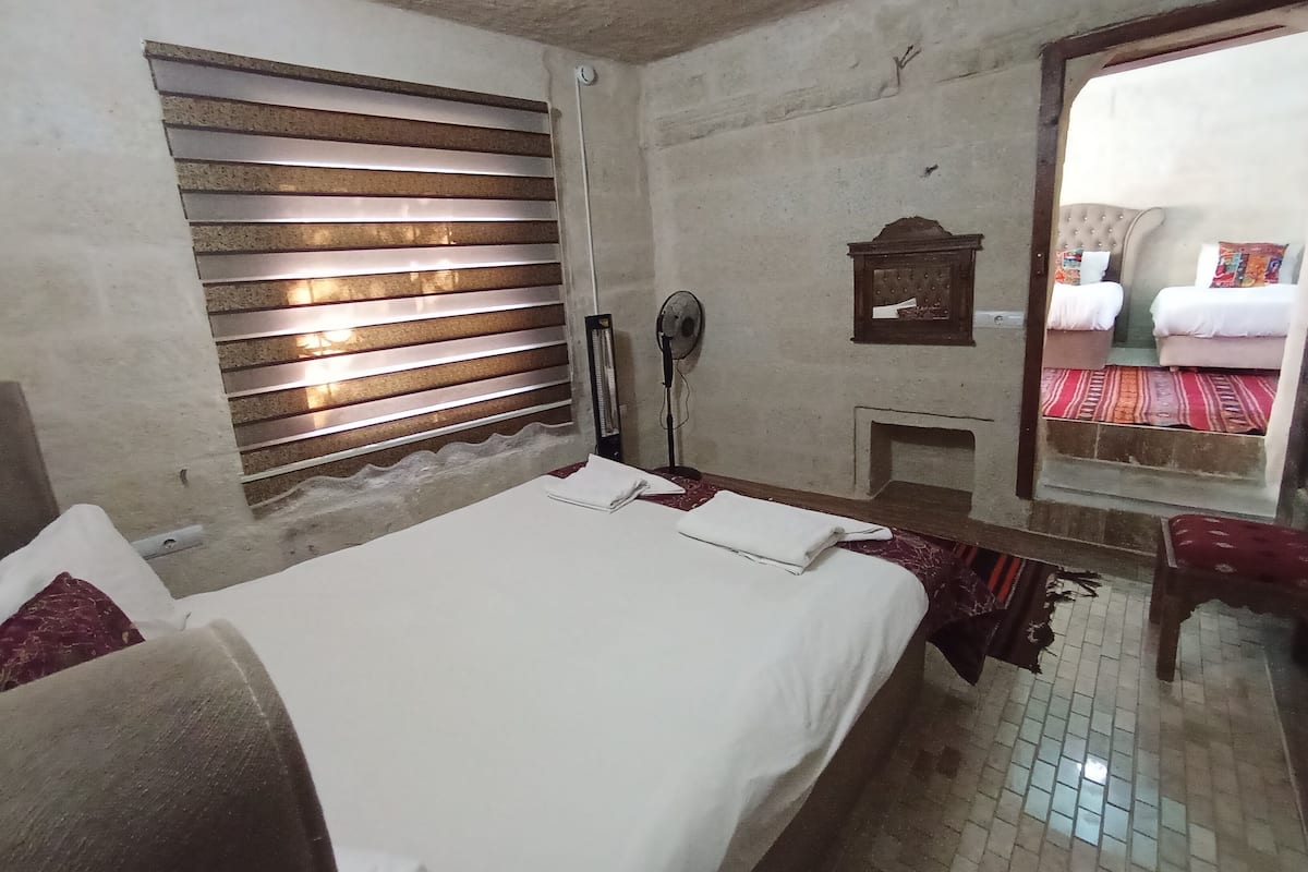 A room with a bed, a fan, a small window with blinds, a mirror, and a view of a balcony with a bed and a rug.
