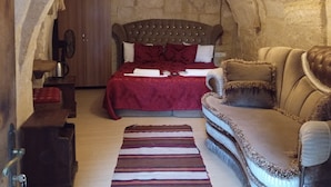 A cozy room with a stone archway, a bed with a red bedspread, a wooden nightstand, a leather sofa, and a striped rug. - Sandik Cave Hotel (Ürgüp)