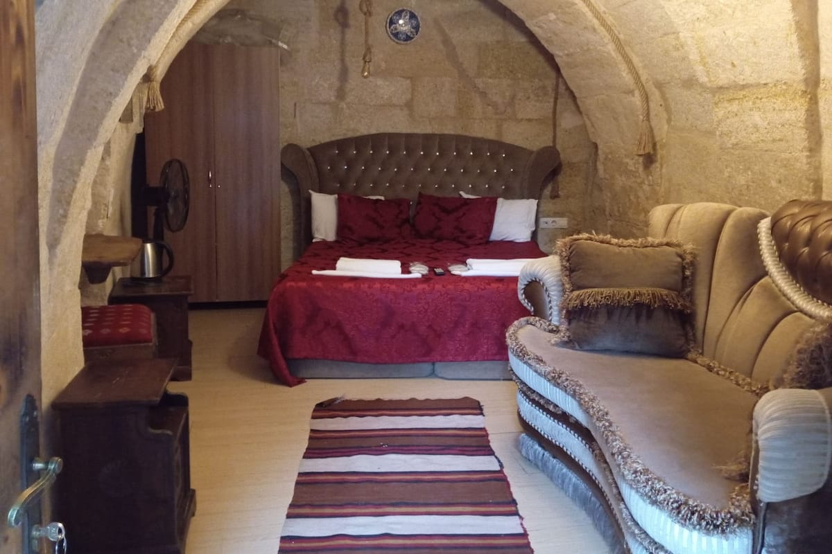 A cozy room with a stone archway, a bed with a red bedspread, a wooden nightstand, a leather sofa, and a striped rug.