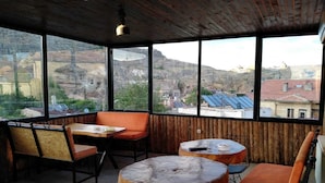 A cozy outdoor seating area with wooden tables and orange cushioned chairs, large windows offering a view of a mountainous landscape and a town below. - Sandik Cave Hotel (Ürgüp)